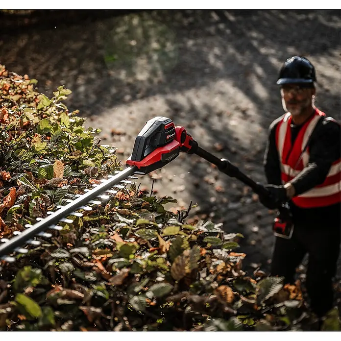 Einhell Professional Teleskop-Heckenschere GP-HH 18/50 Li T BL-Solo - 18 V, Li-Ionen, Anzahl Akkus: Ohne Akku, Schnittlänge: 50 cm - Rote und schwarze Einhell Stiel-Heckenschere mit Metallklingen schneidet eine Hecke, gehalten von einer Person in Sicherheitsweste.