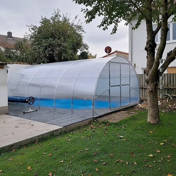 Garten Dream Poolüberdachung Serena, 4mm Polycarbonate - L x B x H: 1.000 x 400 x 230 cm - Eine gewölbte, lichtdurchlässige Poolüberdachung mit silbernem Rahmen bedeckt einen blau ausgekleideten Swimmingpool. Daneben steht eine Poolabdeckrolle.
