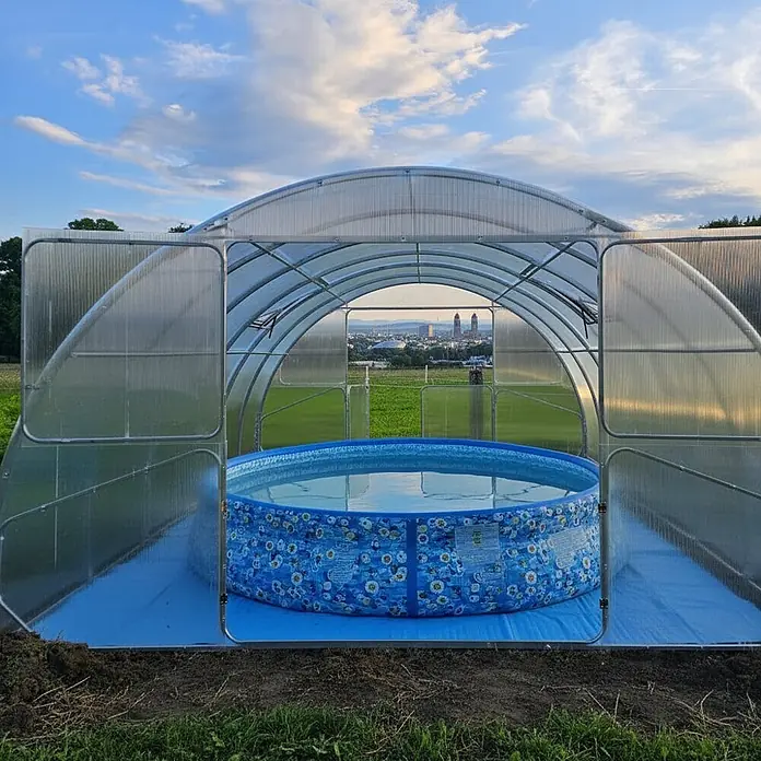 Garten Dream Poolüberdachung Serena, 4mm Polycarbonate - L x B x H: 1.000 x 400 x 230 cm - Ein transparentes Bogengewächshaus umschließt einen blauen aufblasbaren Pool, geöffnet mit Blick auf eine Stadtkulisse.