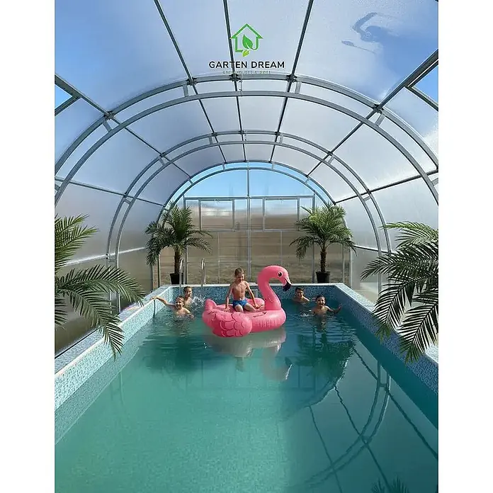 Garten Dream Poolüberdachung Grand Oasis - L x B x H: 600 cm x 400 cm x 2.890 mm - Gewölbte, transparente Schwimmbadüberdachung mit Pool, Palmen und Kindern, die einen rosa Flamingo-Schwimmring nutzen.