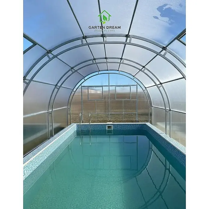 Garten Dream Poolüberdachung Grand Oasis - L x B x H: 600 cm x 400 cm x 2.890 mm - Ein Schwimmbecken mit blauen Fliesen unter einer durchsichtigen, gewölbten Gewächshaus-Überdachung.