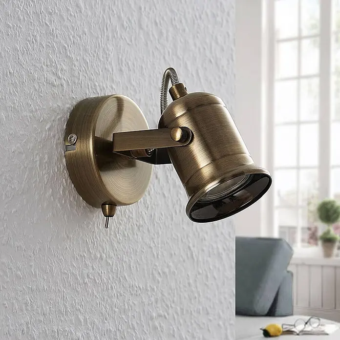 Lindby Wandstrahler Anze - messing / gold, messing / gold, GU10 - Eine verstellbare Wandleuchte aus Antikmessing ist an einer strukturierten weißen Wand in einem Raum mit Fenster montiert.