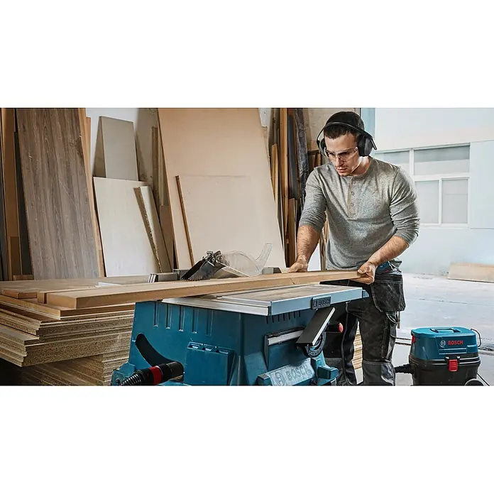 Bosch Professional Tischkreissäge GTS 254 - 1.800 W, Durchmesser Sägeblatt: 250 mm, 4.300 U/min - Mann sägt Holz mit einer blauen Bosch Tischkreissäge, ein blauer Bosch Staubsauger ist für sauberes Arbeiten angeschlossen.