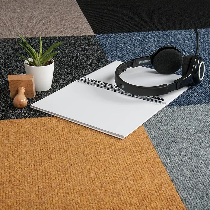 Bricoflor Teppichfliese - Breite: 50 cm, Nadelfilz, Polypropylen, grau - Ein weißes Spiralnotizbuch mit Logitech-Kopfhörern, einer kleinen grünen Pflanze und einem Holzstempel liegt auf einem mehrfarbigen Teppich.