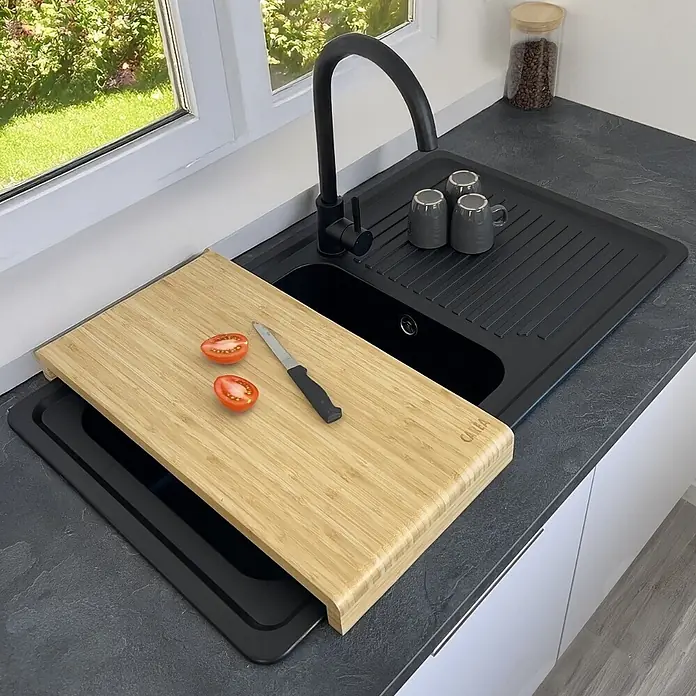 Carea Einbauspüle Venus - 86 x 50 cm, Schwarz, Mit Schneidebrett - Bambus-Schneidebrett mit zwei Tomatenhälften und Messer, auf einem schwarzen Spüleinsatz in einer schwarzen Küchenspüle.