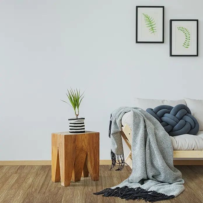 Vinylboden CV-Belag - 500 x 100 cm, Oak Westwood S02 - Heller Holzfuton mit beigem Kissen, grauer gestreifter Decke, dunkelgrauem Knotenpolster. Holztisch mit Topfpflanze und zwei gerahmte Farnbilder an der Wand.