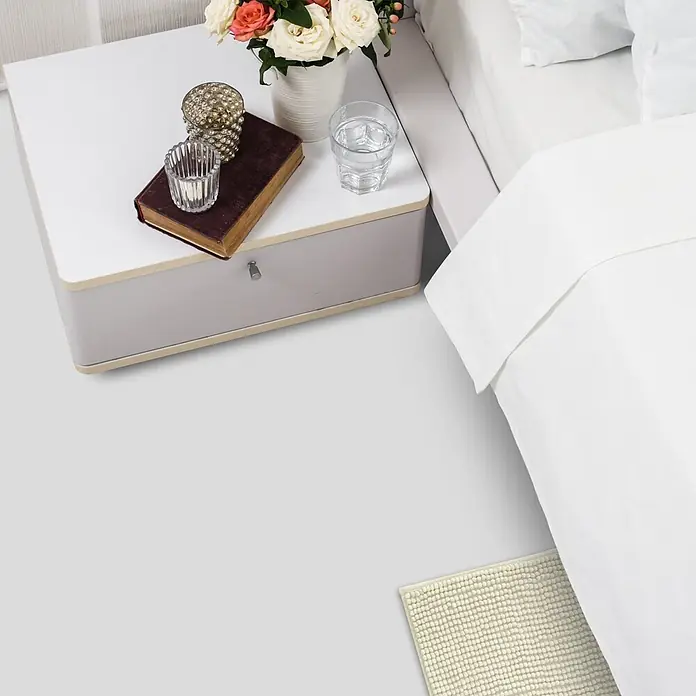 Vinylboden CV-Belag - 500 x 200 cm, Weiß, Expotop - Moderner Nachttisch: weiße Platte, graue Schublade, Blumen, Glas, Buch, Kerzenhalter, neben Bett mit weißer Bettwäsche. Cremefarbener Teppich am Boden.
