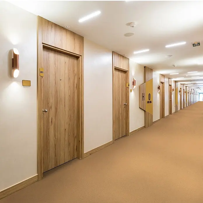 Vinylboden CV-Belag - 50 x 200 cm, Mira 330M - Langer Hotelflur mit Holztüren und warmbraunem Teppich. Moderne Beleuchtung erhellt den Gang.