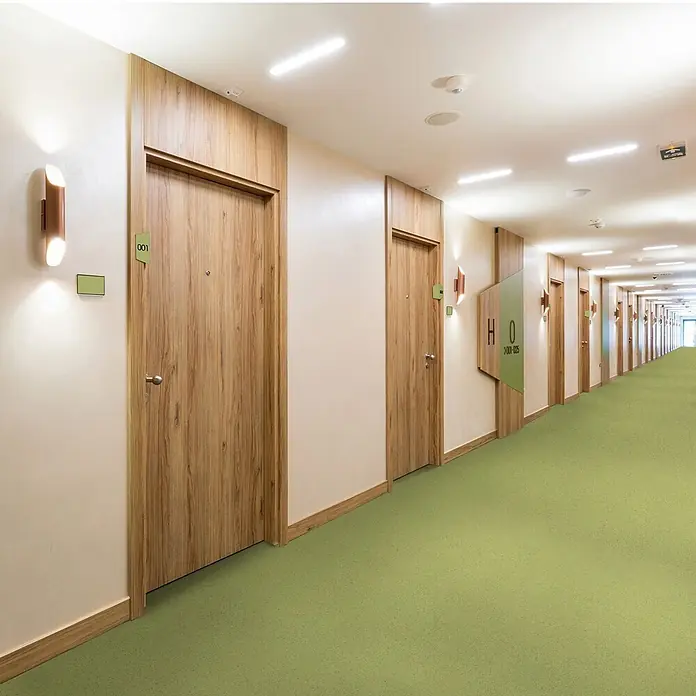 Vinylboden CV-Belag - 150 x 100 cm, Mira 550M - Ein langer Hotelkorridor mit zahlreichen Holztüren, modernen Wandleuchten, Deckenstrahlern und einem hellgrünen Teppich.