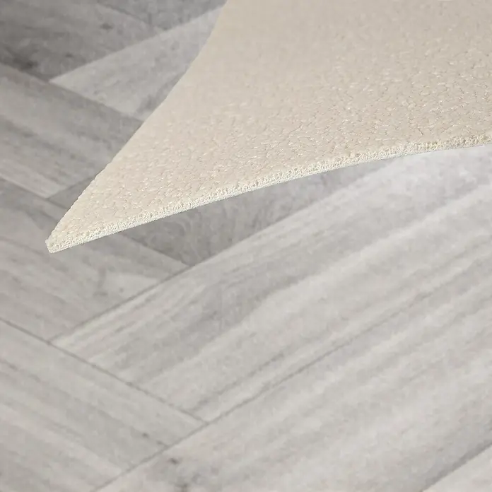 Vinylboden CV-Belag - 400 x 200 cm, Oak Chevron 907M - Hellbeiger, strukturierter Teppich/Matte, Kante angehoben, auf grauem Fischgrätparkett.