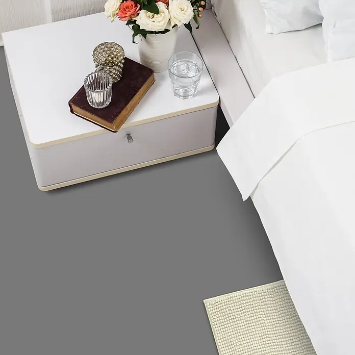 Vinylboden CV-Belag - 2.000 x 200 cm, Grau, Expotop - Weißer Nachttisch mit hellgrauer Schublade, neben einem Bett mit weißer Bettwäsche und einem beigen Teppich.