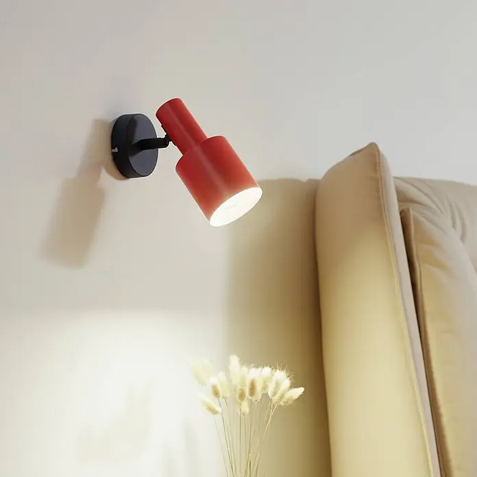Lindby Wandstrahler Ovelia - schwarz, gelb / orange, E27 - Rote Wandleuchte beleuchtet Trockenblumen neben einem beigen Sofa.
