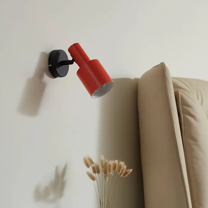 Lindby Wandstrahler Ovelia - schwarz, gelb / orange, E27 - Roter zylindrischer Wandstrahler mit schwarzem Sockel, an einer Wand über getrocknetem Gras, neben einem beigen Sofa montiert.