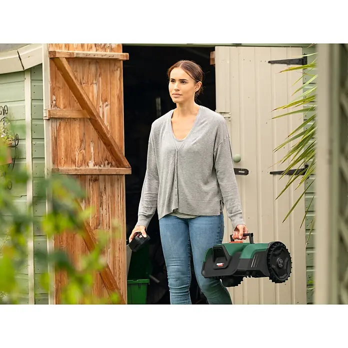 Bosch Power for All 18V Mähroboter VisiMow 18V-200 - 1 Akku, 200 m², Schnittbreite: 18 cm, Kamera-gestützte Navigation, Ultraschallsensorik - Frau trägt grünen und schwarzen Mähroboter und schwarzen Akku aus einem Schuppen, was die Tragbarkeit zeigt.