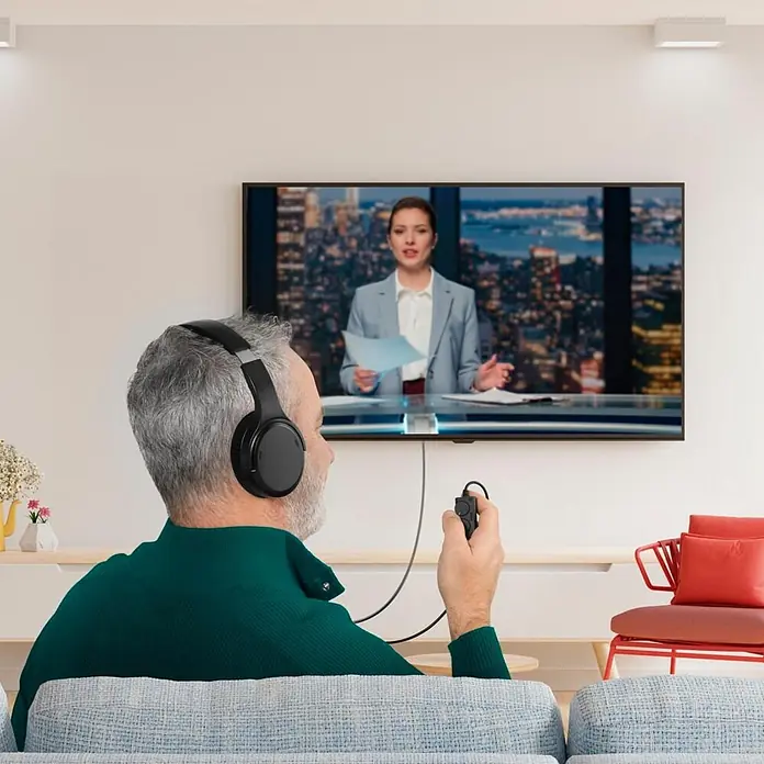 Hama Kopfhörer Fernseh-Kopfhörer Over-Ear langes Kabel - Schwarz - Mann trägt schwarze Kopfhörer, hält ein kleines schwarzes kabelgebundenes Audiogerät und sieht fern.