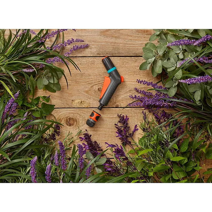 Gardena Classic Kit pistola de limpieza II - Número de formas de rociado: 2 ud., Plástico - Pistola de riego gris y naranja con conector de manguera desconectado sobre fondo de madera, rodeada de plantas verdes y flores moradas.