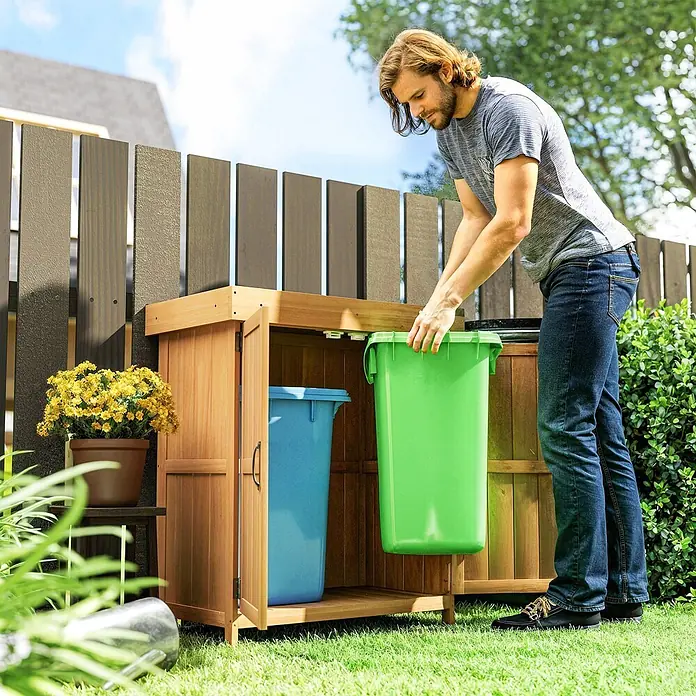 Outsunny Geräteschrank - Außenbreite inkl. Dachüberstand: 74 cm, Außentiefe inkl. Dachüberstand: 43 cm, Holz, Natur - Hölzerner Mülltonnenunterstand mit zwei Recyclingbehältern, ein Mann stellt einen grünen Behälter hinein.
