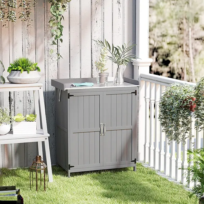 Outsunny Geräteschrank Grau, 74cm x 43cm x 88cm - Außenbreite inkl. Dachüberstand: 74 cm, Außentiefe inkl. Dachüberstand: 43 cm, Holz, Grau - Grauer zweitüriger Outdoor-Aufbewahrungsschrank auf einer Terrasse, umgeben von verschiedenen Grünpflanzen.