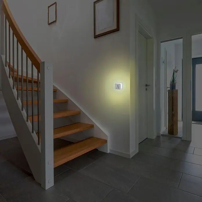 Hama LED-Nachtlicht 00223507 mit Steckdose 2 USB-Ausgänge Bewegungsmelder Lichtsensor -  - Weißes Steckdosen-Nachtlicht erhellt einen dunklen Flur neben einer Holztreppe und spendet sanftes Orientierungslicht.