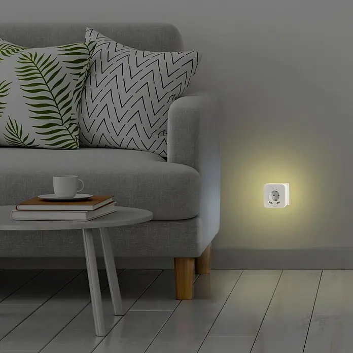 Hama LED-Nachtlicht 00223507 mit Steckdose 2 USB-Ausgänge Bewegungsmelder Lichtsensor -  - Weißer quadratischer Wandstecker mit AC- und USB-Anschlüssen, leuchtet warm als Nachtlicht in einem gedämpften Wohnzimmer.