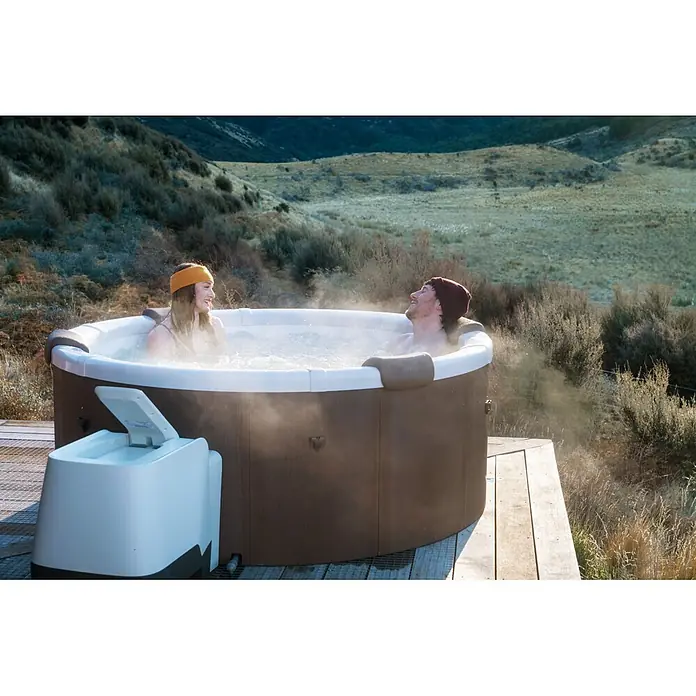 MSpa Mobil-Whirlpool Rowa Frame - Ø x H: 182 x 70 cm, Braun, 970 l - Zwei Personen entspannen sich in einem braun-weißen Whirlpool auf einer Holzterrasse im Freien, Dampf steigt aus dem Wasser.