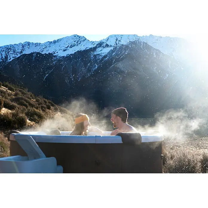 MSpa Mobil-Whirlpool Rowa Frame - Ø x H: 182 x 70 cm, Braun, 970 l - Zwei Personen genießen einen dunkelgrauen Outdoor-Whirlpool mit aufsteigendem Dampf, umgeben von schneebedeckten Bergen.