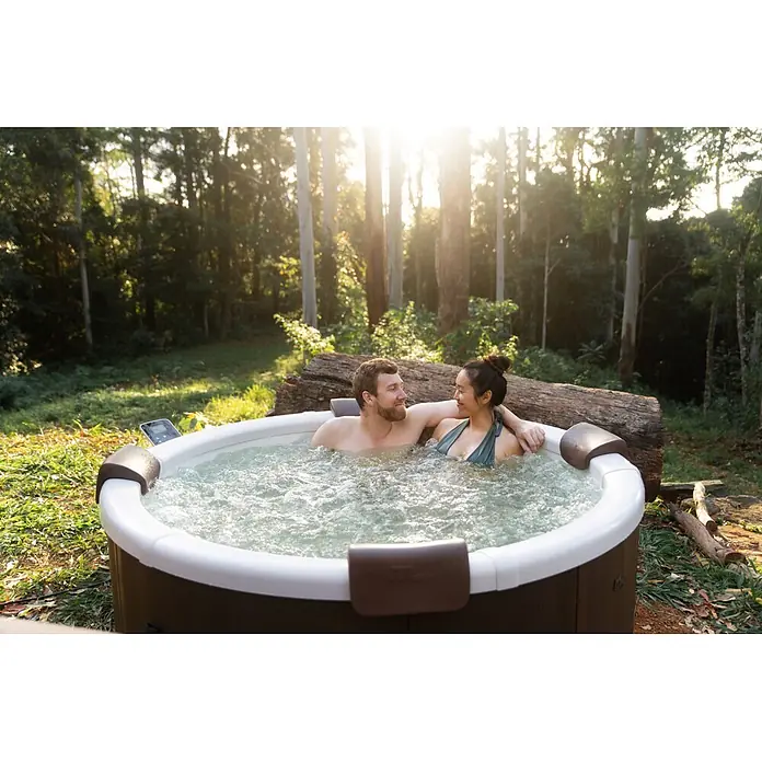 MSpa Mobil-Whirlpool Rowa Frame - Ø x H: 182 x 70 cm, Braun, 970 l - Weißer und brauner Whirlpool mit sprudelndem Wasser, von einem Paar in sonniger Naturkulisse umgeben von Bäumen genutzt.