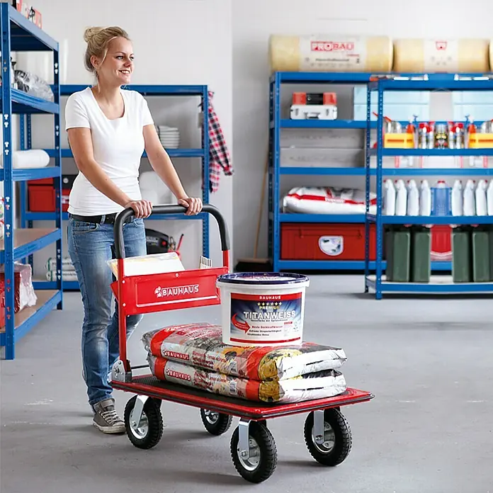 BAUHAUS Carro de plataforma - Ajuste de altura: 83 cm - 100 cm, Acero, 150 kg, Abatible - Mujer empujando un carro rojo con materiales de construcción incluyendo sacos y un cubo de pintura Titanweiss en una ferretería.