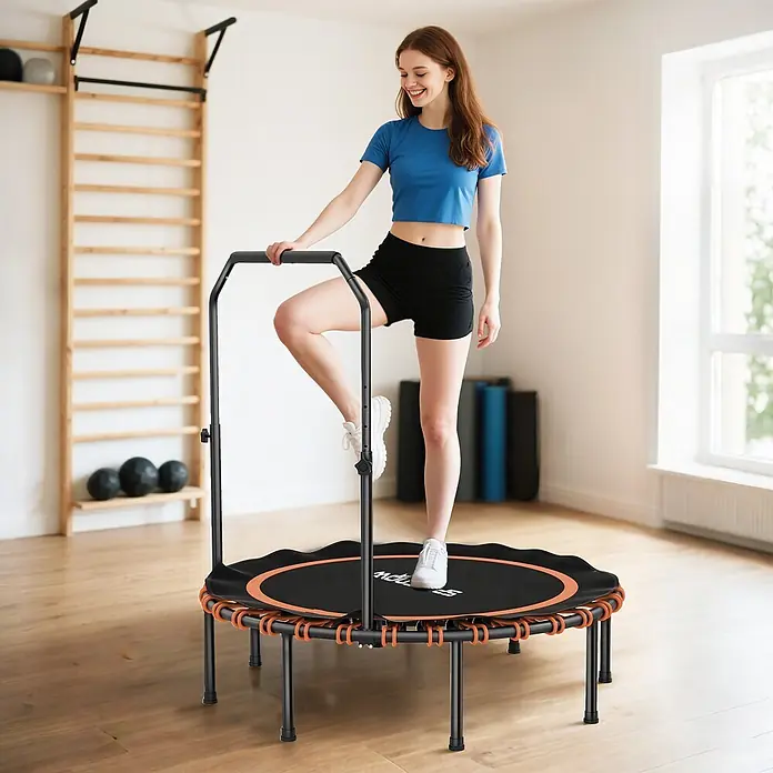 Sportnow Rebounder - Stahl, Schwarz, Orange - Eine Frau lächelt, während sie den verstellbaren Griff eines schwarz-orangen Minitrampolins hält und auf dessen Sprungmatte tritt.