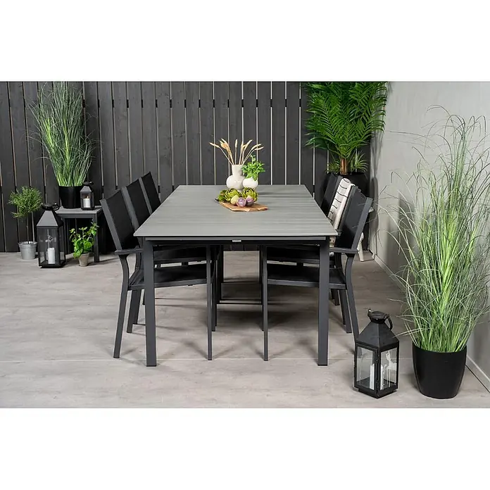 Ebuy24 Gartentisch-Set Levels - Gesamtumfang: 7 -tlg., L x B x H: 229 x 100 x 74 cm, Ø x H: 229 x 74 cm, Aintwood, Stapelbar - Grauer Esstisch für den Außenbereich mit sechs dunkelgrauen Stühlen auf einer Terrasse mit Zierpflanzen und Laternen.
