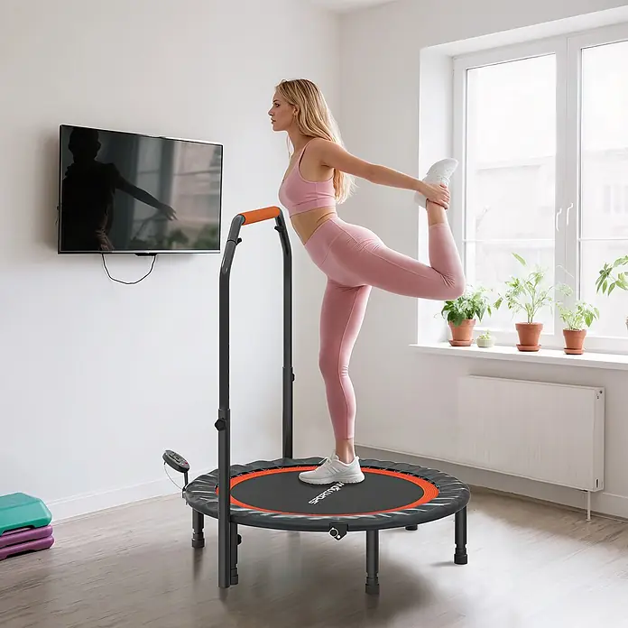 Sportnow Rebounder - Stahl, Schwarz - Eine Frau in rosa Sportkleidung dehnt sich auf einem schwarzen und orangefarbenen Minitrampolin mit Griff in einem hellen Raum.