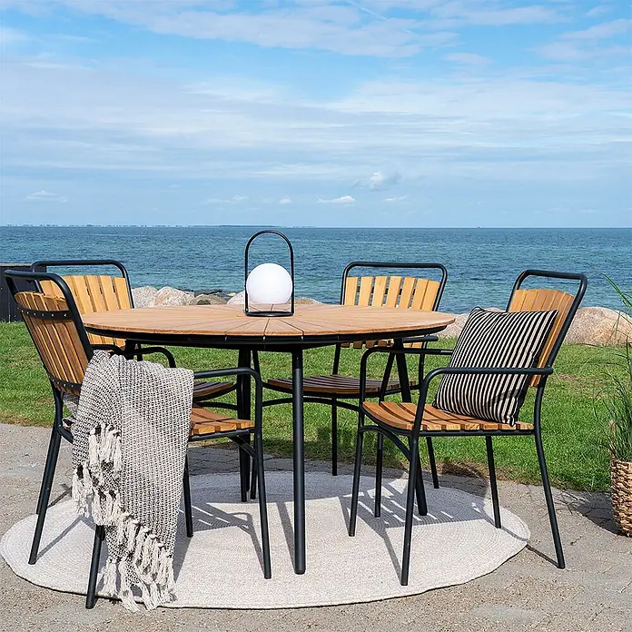 Ebuy24 Gartentisch-Set Cleveland - Gesamtumfang: 5 -tlg., L x B x H: 130 x 130 x 74 cm, Ø x H: 130 x 74 cm, Teak Metall Aluminum Polyester, Stapelbar - Runde Outdoor-Essgruppe aus Holz mit schwarz gerahmten Stühlen und Accessoires auf einem Teppich am Meer.
