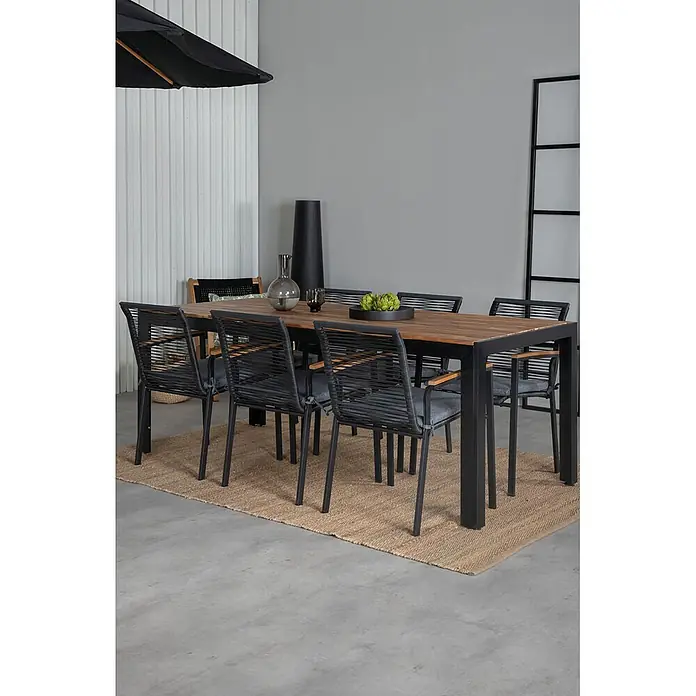 Ebuy24 Gartentisch-Set Bois - Gesamtumfang: 7 -tlg., L x B x H: 205 x 90 x 76 cm, Ø x H: 90 x 76 cm, 100 % Polyester, Holz, Rattan, Stahl Akazie, Aluminium, Stapelbar - Ein Outdoor-Esstisch-Set mit rechteckigem Holztisch und sechs schwarzen geflochtenen Stühlen mit grauen Kissen auf einem Juteteppich.