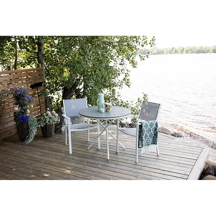 Ebuy24 Gartentisch-Set Parma - Gesamtumfang: 7 -tlg., L x B x H: 90 x 90 x 74 cm, Ø x H: 90 x 74 cm, Textil Nonwood, Stapelbar - Weißes Outdoor-Essset mit hellgrauem Tisch und Stühlen auf einer Terrasse am See.