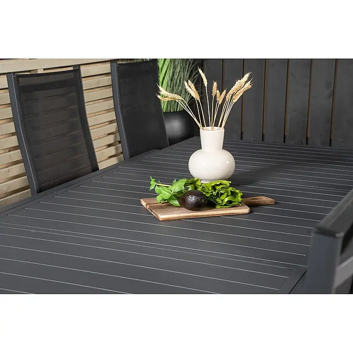 Ebuy24 Gartentisch-Set Marbella - Gesamtumfang: 9 -tlg., L x B x H: 100 x 160 x 75 cm, Ø x H: 100 x 75 cm, 30 % Polyester, 70 % PVC, Aluminium, Textil Aluminium, Stapelbar - Dunkelgrauer Lamellen-Gartentisch mit passenden Stühlen in einer Gartenumgebung.