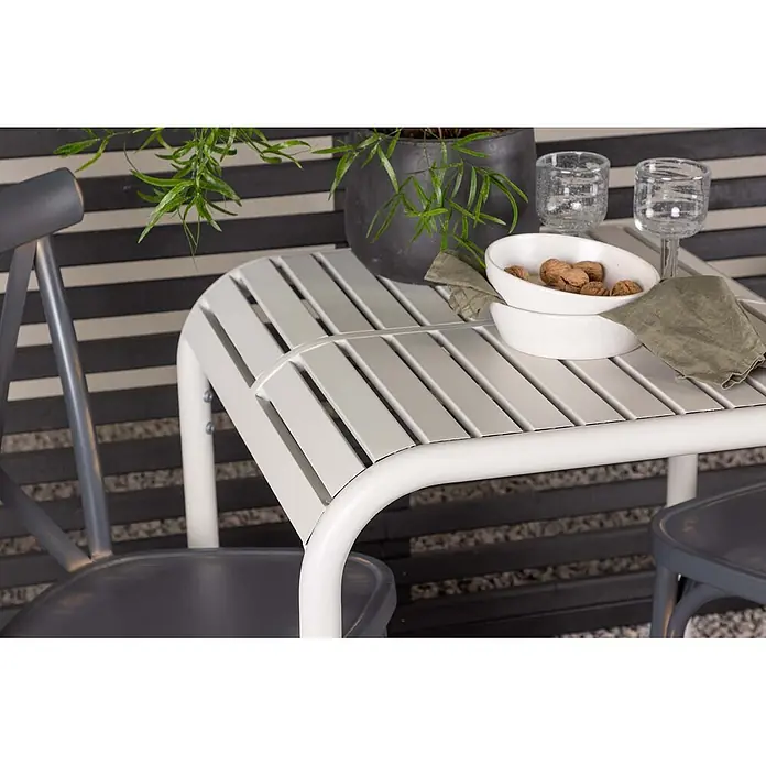 Ebuy24 Gartentisch-Set Borneo - Gesamtumfang: 3 -tlg., L x B x H: 70 x 70 x 75 cm, Ø x H: 70 x 75 cm, Aluminium, Stapelbar - Ein hellgrauer Lamellen-Outdoor-Tisch mit weißen gebogenen Beinen, mit Schalen und Gläsern neben dunkelgrauen Stühlen.