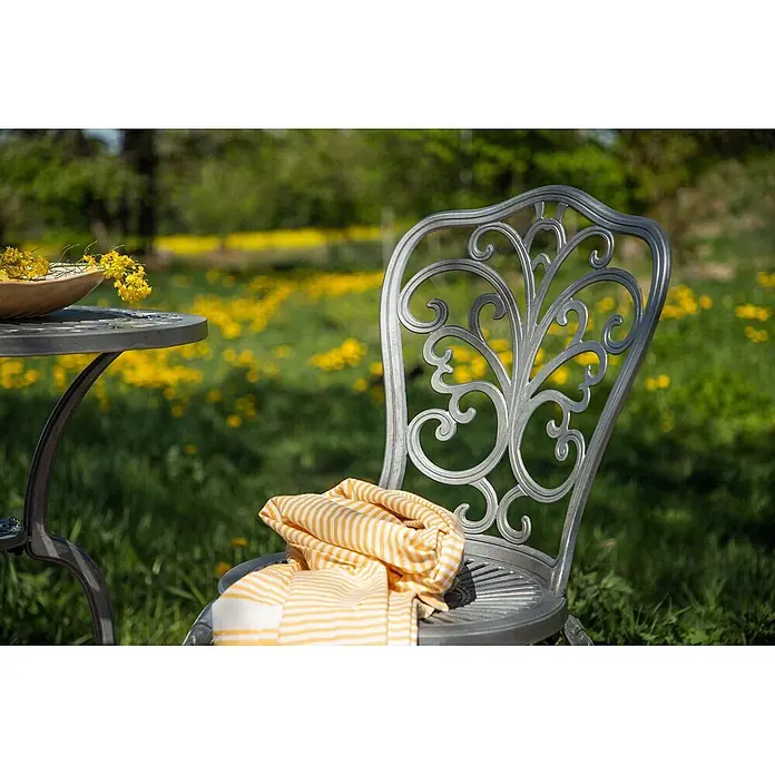 Ebuy24 Gartentisch-Set Nandin - Gesamtumfang: 3 -tlg., L x B x H: 60 x 60 x 73 cm, Ø x H: 60 x 73 cm, Aluminium, Stapelbar - Silberner Zier-Metall-Gartenstuhl und -tisch mit gelb gestreiftem Handtuch, im Freien aufgestellt.