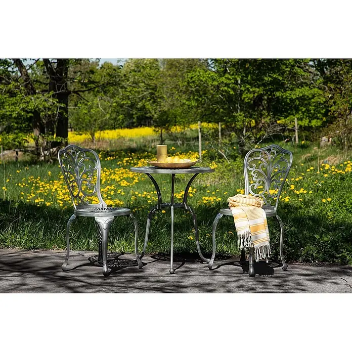 Ebuy24 Gartentisch-Set Nandin - Gesamtumfang: 3 -tlg., L x B x H: 60 x 60 x 73 cm, Ø x H: 60 x 73 cm, Aluminium, Stapelbar - Silbergraues Metall-Bistro-Set mit 2 Stühlen und Tisch im Freien. Gelbe Früchte/Tassen auf dem Tisch, gestreiftes Handtuch auf einem Stuhl.