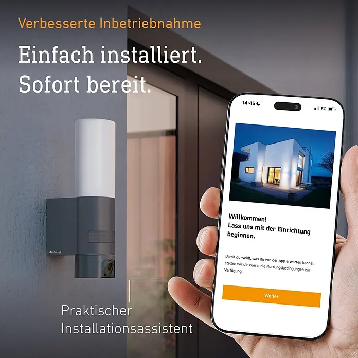 Steinel LED-Sensor-Außenwandleuchte L 625 CAM SC - 14 W, L x B x H: 13,1 x 7,8 x 30,5 cm, Anthrazit/Weiß, Warmweiß - Eine dunkelgraue und weiße Außenleuchte an einer Wand, daneben ein Smartphone mit der Einrichtungs-App in einer Hand.