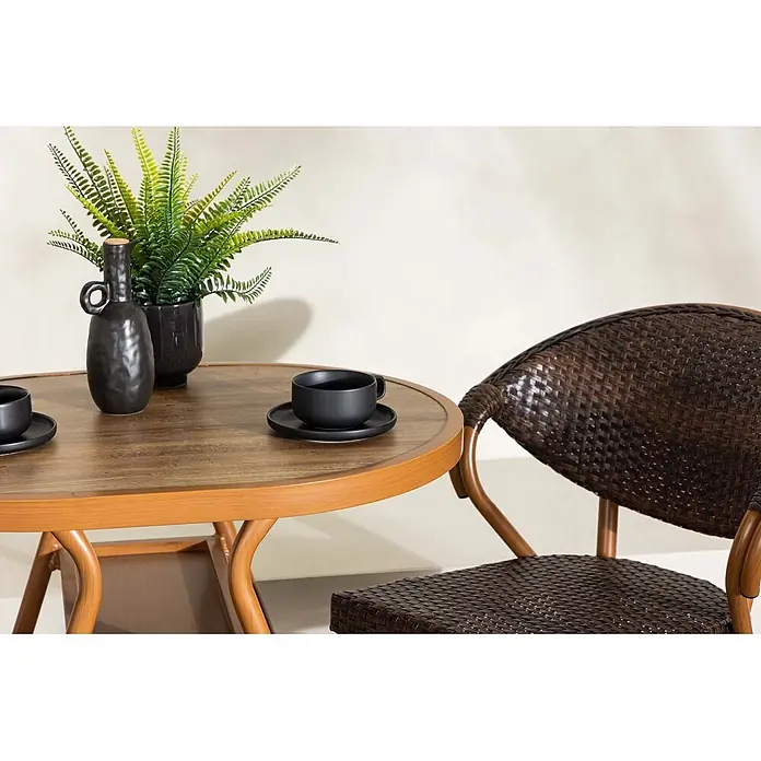 Ebuy24 Gartentisch-Set Abrantes - Gesamtumfang: 3 -tlg., L x B x H: 80 x 80 x 74 cm, Ø x H: 80 x 74 cm, Aluminium, Stapelbar - Runder Holztisch mit hellbraunem Gestell, einer schwarzen Tasse und einer Pflanze, neben einem dunkelbraunen Korbstuhl.