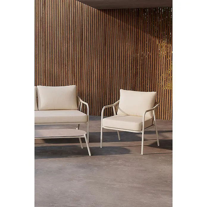 Ebuy24 Loungemöbel-Set Sicily - 4 -tlg., Aluminium, Beige - Outdoor-Sitzgruppe: ein Sofa und Sessel mit weißem Rahmen und cremefarbenen Kissen auf einer Betonterrasse vor einer Holzlamellenwand.
