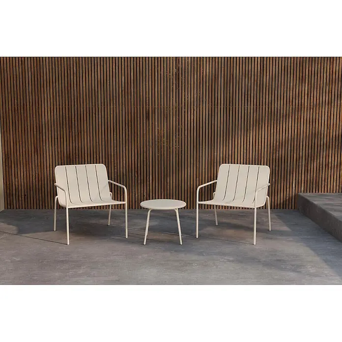 Ebuy24 Loungemöbel-Set Minos - 3 -tlg., Stahl, Beige - Zwei cremefarbene Metall-Gartenstühle und ein passender kleiner Tisch auf einer Betonterrasse vor einer Holzlattenwand.