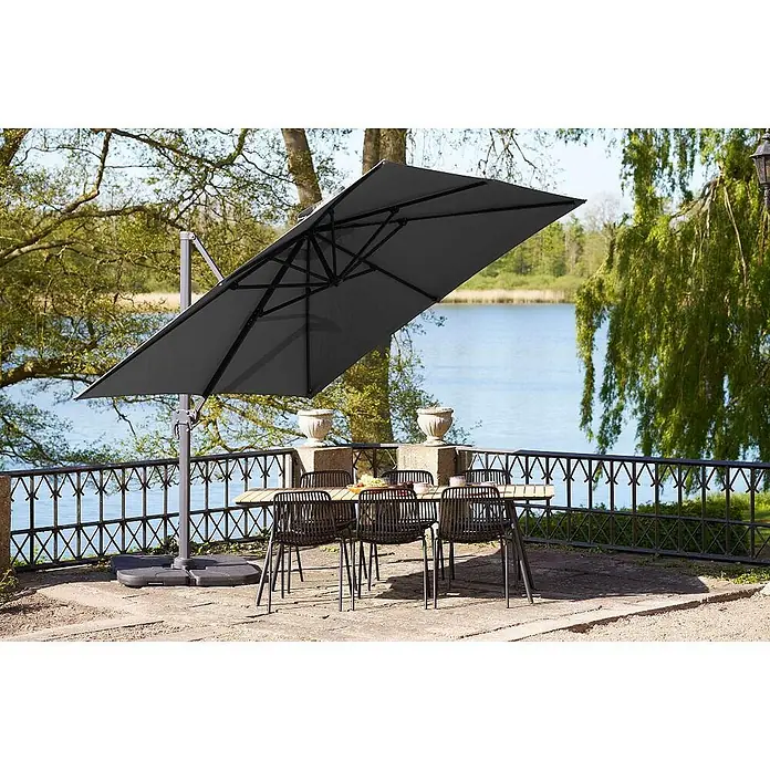 Ebuy24 Marktschirm Bea - Schwarz - Ein großer grauer, rechteckiger Freiarmschirm beschattet einen Esstisch mit Stühlen auf einer Terrasse am See.