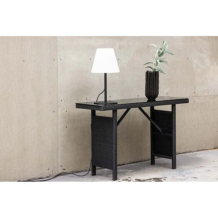 Ebuy24 Garten-Beistelltisch Padova - L x B x H: 40 x 120 x 74 cm, Korbmöbel, Stapelbar, Sonstige, Schwarz - Schwarzer Rattan-Bartisch mit Glasplatte, darauf eine weiße Lampe und eine schwarze Vase mit Grünzeug.