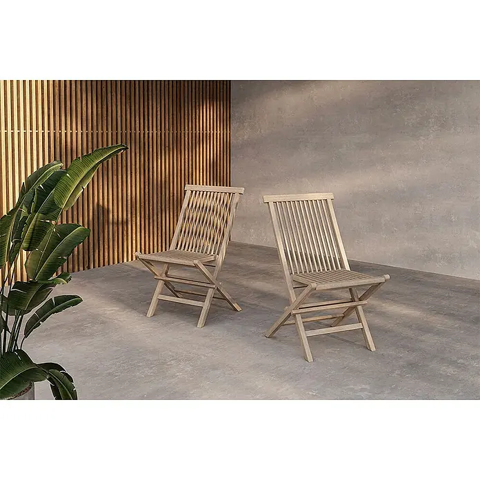 Ebuy24 Gartenstuhl Kenya 2er-Set - Teak, Whitewash, B x T x H: 47 x 62 x 89 cm - Zwei Klappstühle aus Naturholz stehen im Freien auf einem Betonboden neben einer Pflanze.