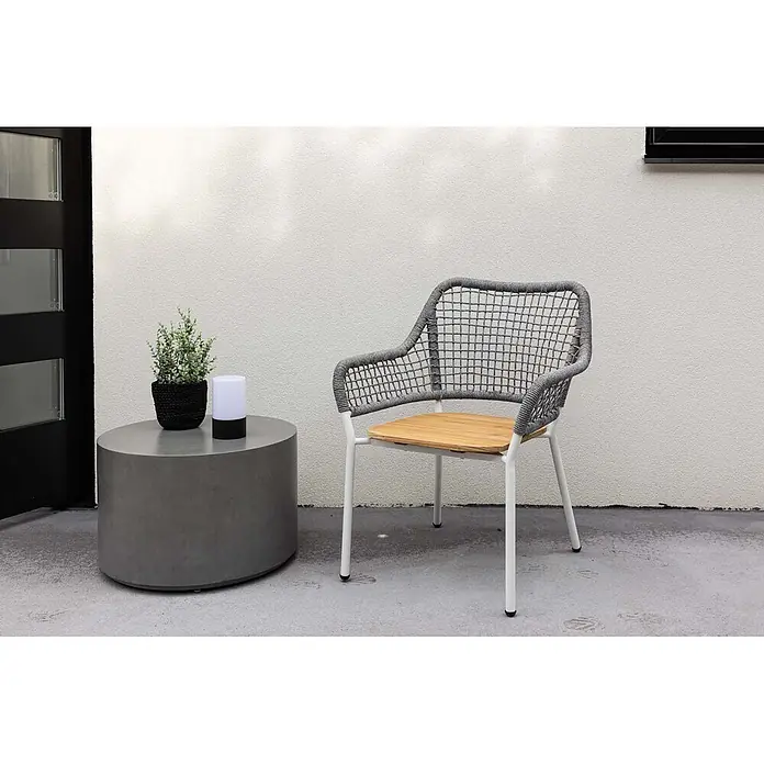 Ebuy24 Gartensessel Amora - 1 Stk., Teak, Grau, B x T x H: 59 x 55 x 88 cm - Ein runder Beistelltisch aus grauem Beton neben einem Outdoor-Stuhl mit grauem Geflecht, Holzplatte und weißen Beinen, auf einer Terrasse.