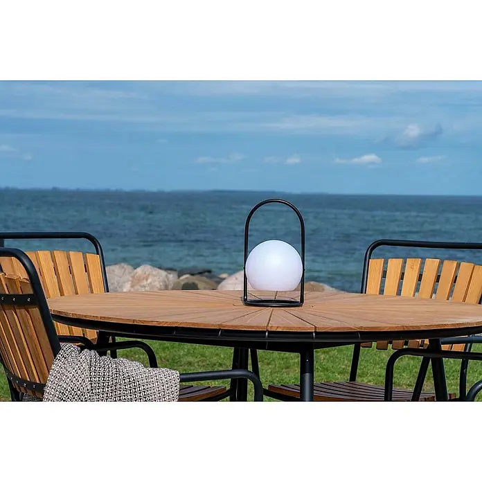 Ebuy24 Gartenstuhl Cleveland 4er-Set - Teakholz Metall, Teak, B x T x H: 64 x 58 x 91 cm - Weiße kugelförmige Außenleuchte mit schwarzem Griff auf einem Holztisch am Meer.