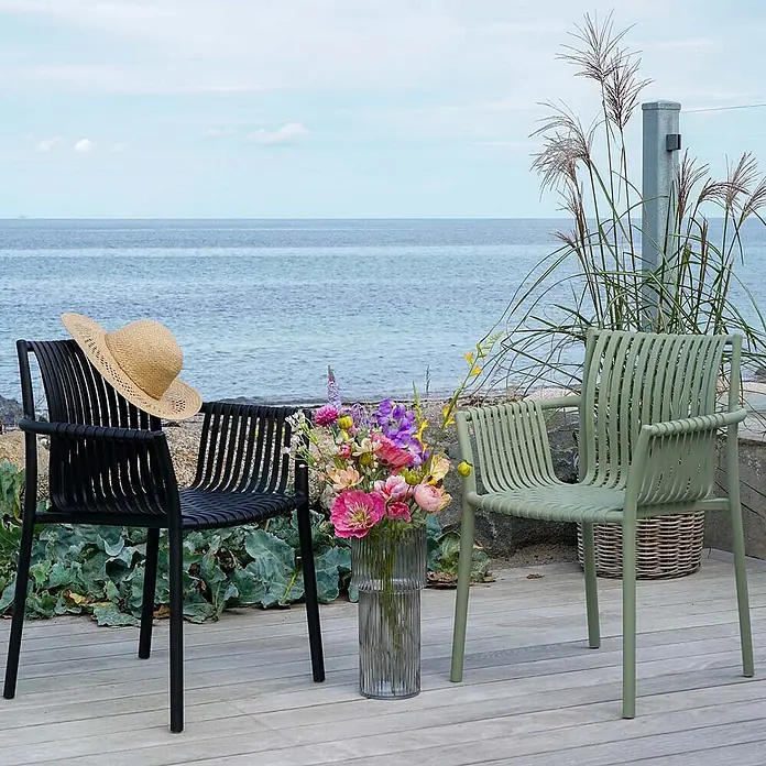 Ebuy24 Gartenstuhl Tulsa 4er-Set - Polypropylen, Schwarz, B x T x H: 57 x 60 x 83 cm - Zwei gerippte Outdoor-Stühle, ein schwarzer mit Strohhut und ein salbeigrüner, auf einer Holzterrasse am Meer mit Blumenvase.