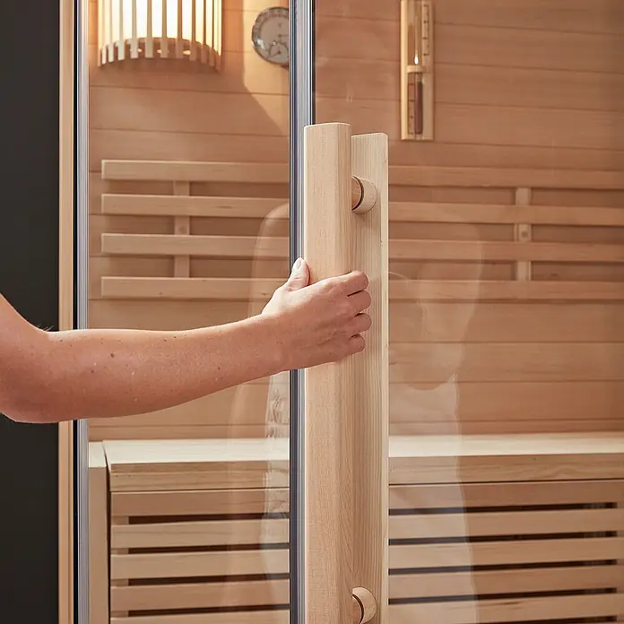 Home Deluxe Elementsauna SKYLINE XL - Elektroofen, Steuerung Ofen: Integriert - Hand öffnet eine Saunatür mit natürlichem Holzgriff, die das helle Holzinterieur zeigt.