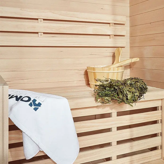 Home Deluxe Elementsauna SKYLINE XL - Elektroofen, Steuerung Ofen: Integriert - Helle Holzsauna mit Bank, Holzeimer, Kelle, Wenik und weißem Handtuch mit blauem Logo.